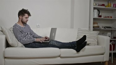 Jaunas vyras, dirbantis nešiojamajame kompiuteryje, sėdi ant sofos