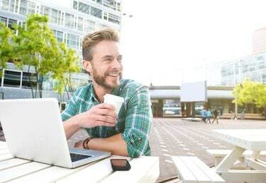 איש מחייך יושב בחוץ עם מחשב נייד וקפה