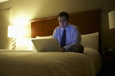 Affärsman sitter med benen över kors på sängen i hotellrum och arbetar på bärbar dator